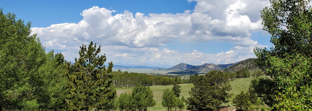 Image of the Front Range in Colorado under a blue sky with white puffy clouds casting shadows across the ground and green trees.